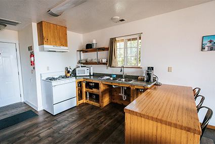 View from the kitchen. A couch and arm chair are in the living room. There are some large doors beyond the living room leading to the outside.